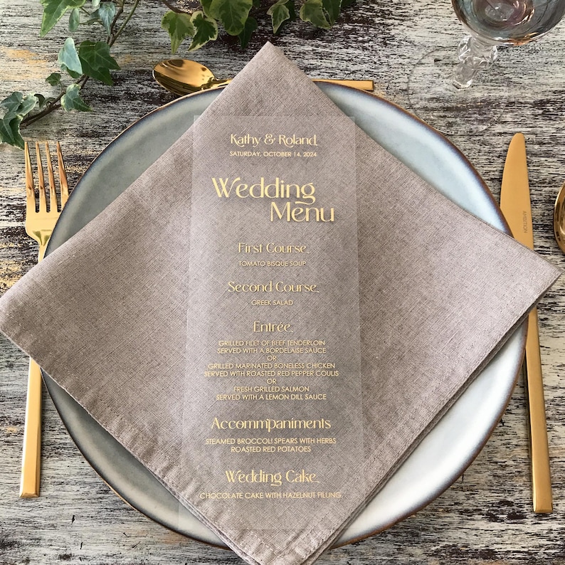Op de afbeelding: Een helder acryl trouwmenu met gouden letters, met gerechten zoals tomatensoep en gegrilde zalm. Het menu ligt op een gevouwen beige linnen servet, op een grijze plaat met gouden bestek. Het menu is gepersonaliseerd voor Kathy & Roland.