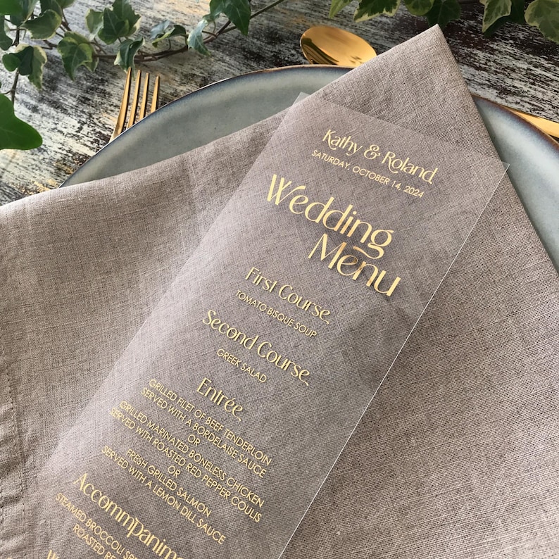 Op de afbeelding: Een heldere acryl trouwmenu met gouden letters, met de namen "Kathy & Roland" en de datum "Zaterdag 14 oktober 2024." Het menu bevat gerechten zoals tomatensoep, Griekse salade en diverse hoofdgerechten. Het ligt op een beige linnen servet.