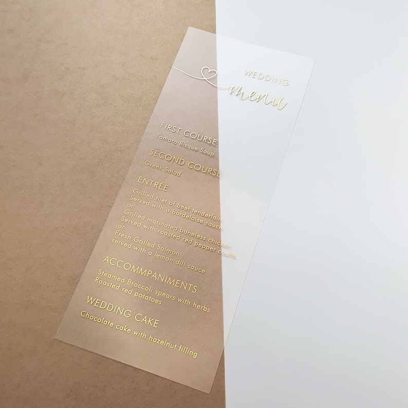 May include: A clear acrylic wedding menu card with gold lettering. The menu includes a first course of tomato bisque soup, a second course of Greek salad, an entree of grilled filet of beef tenderloin or grilled marinated boneless chicken, and a wedding cake with hazelnut filling.