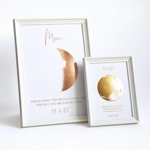 May include: Two white-framed prints featuring metallic moon illustrations. The larger print says "Mom" with a copper crescent moon and "THIS IS WHAT THE MOON LOOKED LIKE THE DAY YOU BECAME MY MOM 19.4.82". The smaller print says "DAD" with a gold full moon.