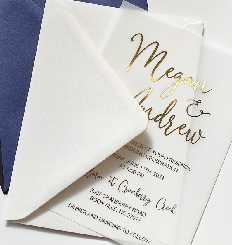 May include: A clear acrylic wedding invitation with gold foil lettering. The invitation reads "Megan & Andrew" and includes the wedding date, time, and location. The invitation is on a white envelope.