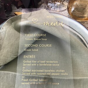 May include: A clear acrylic wedding menu card with gold lettering. The menu includes a first course of Tomato Bisque Soup, a second course of Greek Salad, and an entree choice of Grilled Filet of Beef Tenderloin, Grilled Marinated Boneless Chicken, or Fresh Grilled Salmon.