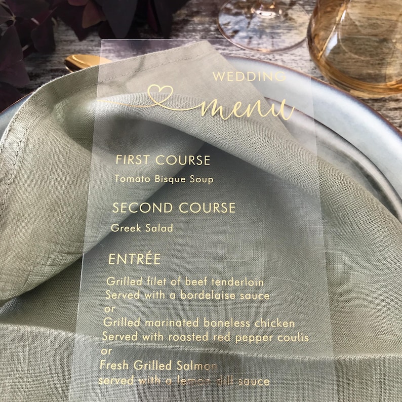 May include: A clear acrylic wedding menu card with gold lettering. The menu includes a first course of Tomato Bisque Soup, a second course of Greek Salad, and an entree choice of Grilled Filet of Beef Tenderloin, Grilled Marinated Boneless Chicken, or Fresh Grilled Salmon.