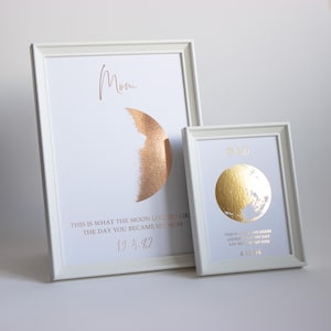 May include: Two framed prints with white frames. One features a copper-colored half-moon design with the word "Mom" and the text "This is what the moon looked like the day you became my Mom" and the date 19.4.82. The other has a gold moon design with the word "Dad" and the date 4.13.04.
