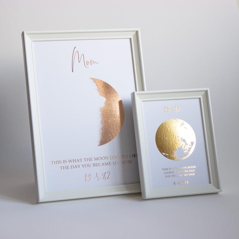 May include: Two framed prints with white frames. One features a copper-colored half-moon design with the word "Mom" and the text "This is what the moon looked like the day you became my Mom" and the date 19.4.82. The other has a gold moon design with the word "Dad" and the date 4.13.04.