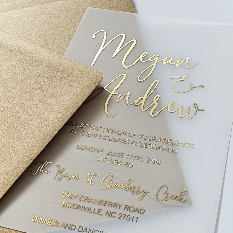 May include: A clear acrylic wedding invitation with gold foil lettering. The invitation reads "Megan & Andrew" and includes the details of their wedding celebration at "The Barn at Cranberry Creek" on Sunday, 17th June 2024 at 5:00 PM. The address is 2907 Cranberry Road, Boonville, NC 27011.