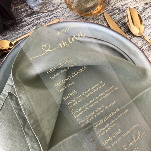 May include: A clear acrylic wedding menu card with gold lettering. The menu includes the courses: First Course, Tomato Bisque Soup; Second Course, Greek Salad; Entr&eacute;e, Grilled Filet of Beef Tenderloin, Grilled Marinated Boneless Chicken, Fresh Grilled Salmon; Accompaniments, Steamed Broccoli, Roasted Red Potatoes; Wedding Cake. The card also includes the text "Wedding Menu" and "& michael 2024".