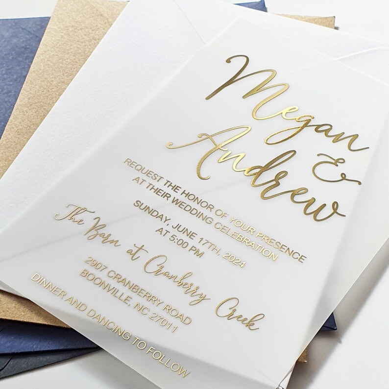 May include: A white wedding invitation with gold lettering. The invitation reads "Megan & Andrew" and includes the date, time, and location of the wedding. The text reads "Request the honour of your presence at their wedding celebration. Sunday, June 17th, 2024 at 5:00 PM. The Barn at Cranberry Creek, 2907 Cranberry Road, Boonville, NC 27011. Dinner and dancing to follow."