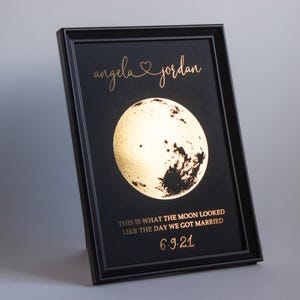 May include: Black framed print featuring a gold foil moon illustration. The text reads "angela & jordan" above the moon, with the phrase "THIS IS WHAT THE MOON LOOKED LIKE THE DAY WE GOT MARRIED" and the date "6.9.21" below.