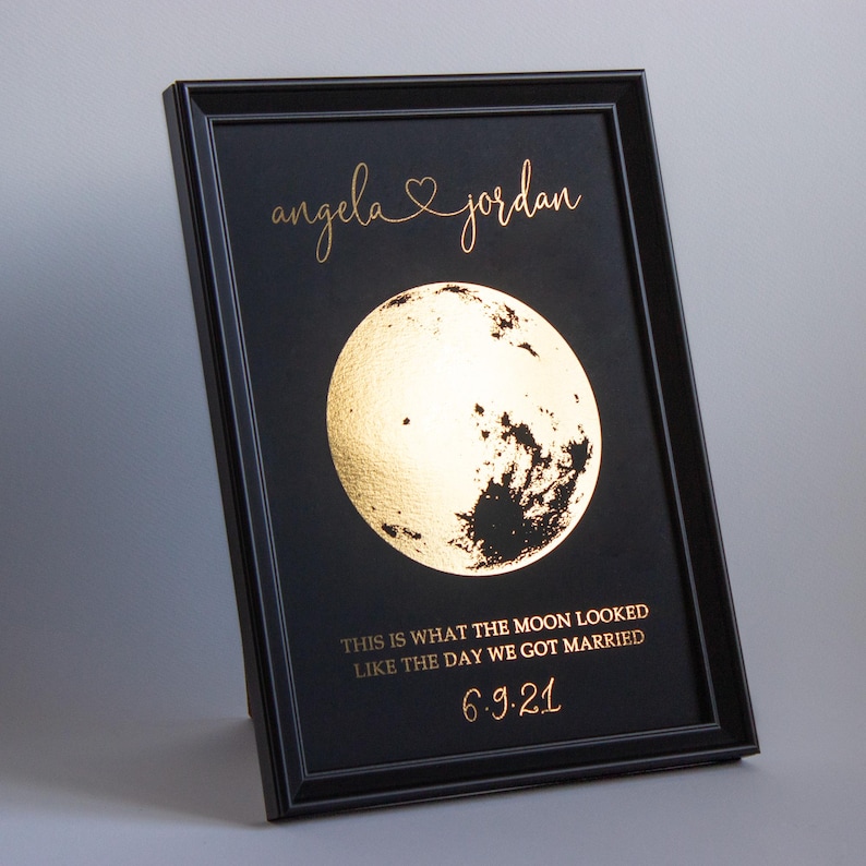 May include: Black framed print featuring a gold foil moon illustration. The text reads "angela & jordan" above the moon, with the phrase "THIS IS WHAT THE MOON LOOKED LIKE THE DAY WE GOT MARRIED" and the date "6.9.21" below.