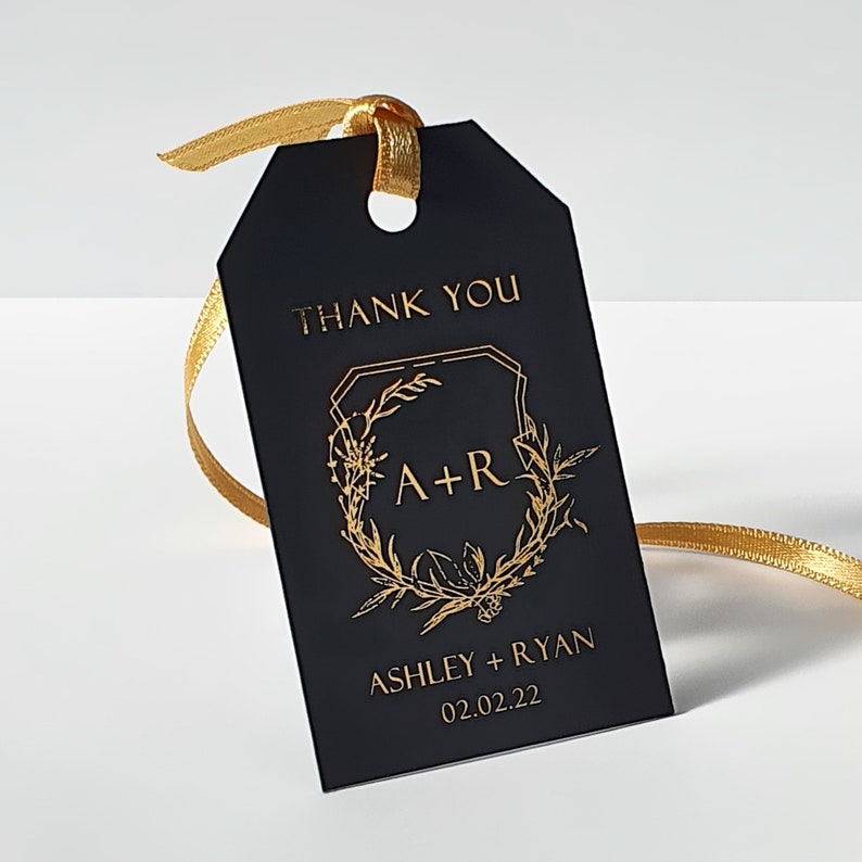 May include: A rectangular black gift tag featuring gold foil text that reads THANK YOU, A+R, ASHLEY + RYAN, and 02.02.22. The tag includes a gold botanical wreath graphic and a gold ribbon threaded through a hole at the top of the tag.