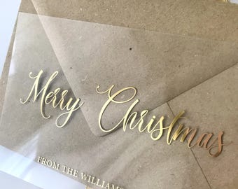 May include: A kraft paper gift bag with a clear overlay. The words "Merry Christmas" are in gold script across the front. Below, the text reads "FROM THE WILLIAMS FAMILY" in a smaller gold font. A gold ribbon is attached.