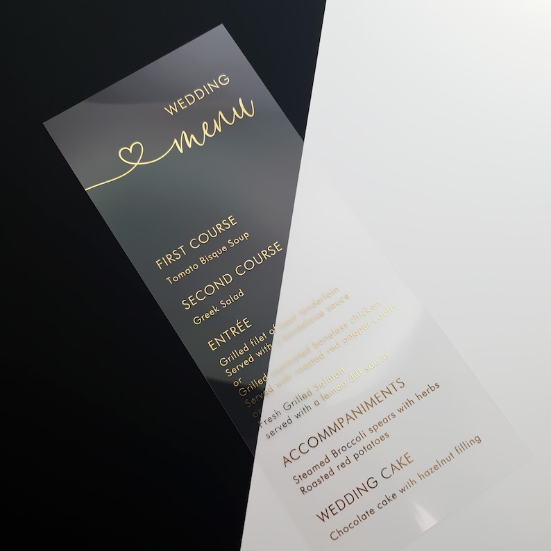 May include: A transparent wedding menu card with gold lettering. The menu includes Tomato Bisque Soup, Greek Salad, Grilled Filet of Salmon, Steamed Broccoli, Roasted Red Potatoes, and Chocolate Cake with Hazelnut Filling.