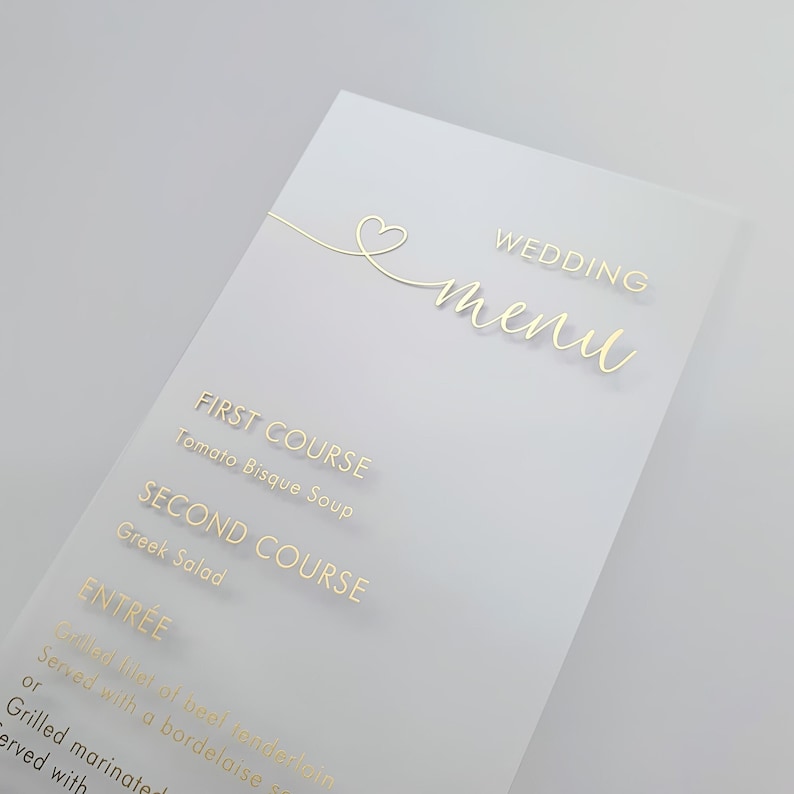 May include: A white wedding menu card with gold lettering. The menu includes: First Course - Tomato Bisque Soup, Second Course - Greek Salad, Entr&eacute;e - Grilled filet of beef tenderloin served with a bordelaise or grilled marinated served with.