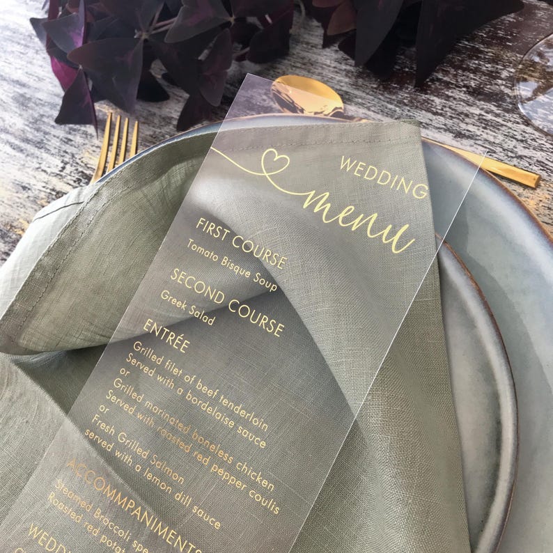May include: A clear acrylic wedding menu card with gold lettering. The menu includes the following courses: First Course - Tomato Bisque Soup, Second Course - Greek Salad, Entr&eacute;e - Grilled filet of beef tenderloin served with a bordelaise sauce or grilled marinated boneless chicken served with roasted red pepper coulis or fresh grilled salmon served with a lemon dill sauce, Accompaniments - Steamed Broccoli, Roasted red potato.