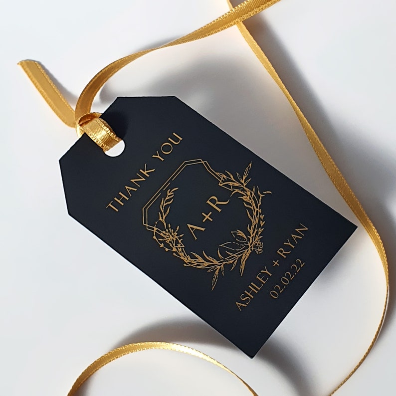 May include: A rectangular black gift tag with gold foil lettering and a gold ribbon. The tag features the text THANK YOU, a floral wreath surrounding the initials A+R, and the names ASHLEY + RYAN with the date 02.02.22 printed in gold.