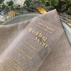 Op de afbeelding: Een heldere trouwmenukaart met gouden letters, met de namen "Kathy & Roland" en de datum "Zaterdag 14 oktober 2024." Het menu bevat gerechten zoals tomatensoep en Griekse salade, liggend op een linnen servet.