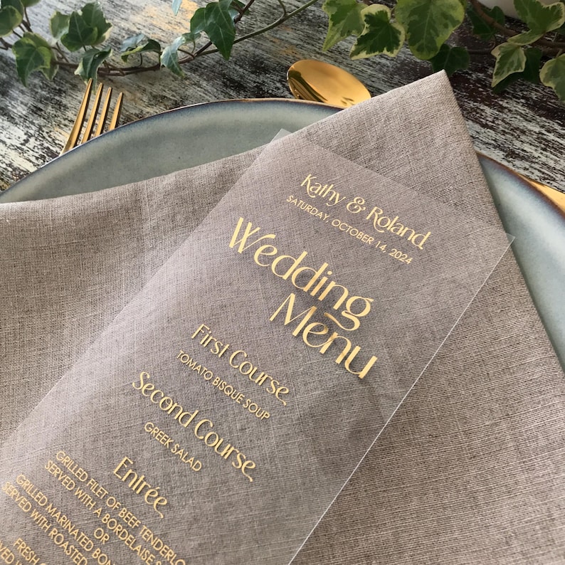 Op de afbeelding: Een heldere trouwmenukaart met gouden letters, met de namen "Kathy & Roland" en de datum "Zaterdag 14 oktober 2024." Het menu bevat gerechten zoals tomatensoep en Griekse salade, liggend op een linnen servet.