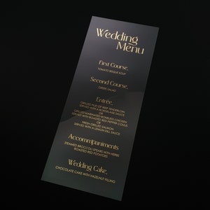 Op de afbeelding: Een zwart trouwmenu met gouden letters. Het menu bevat gerechten zoals tomatenbisque soep, Griekse salade en gegrilde ossenhaas. Het menu bevat ook de tekst "Trouwmenu".
