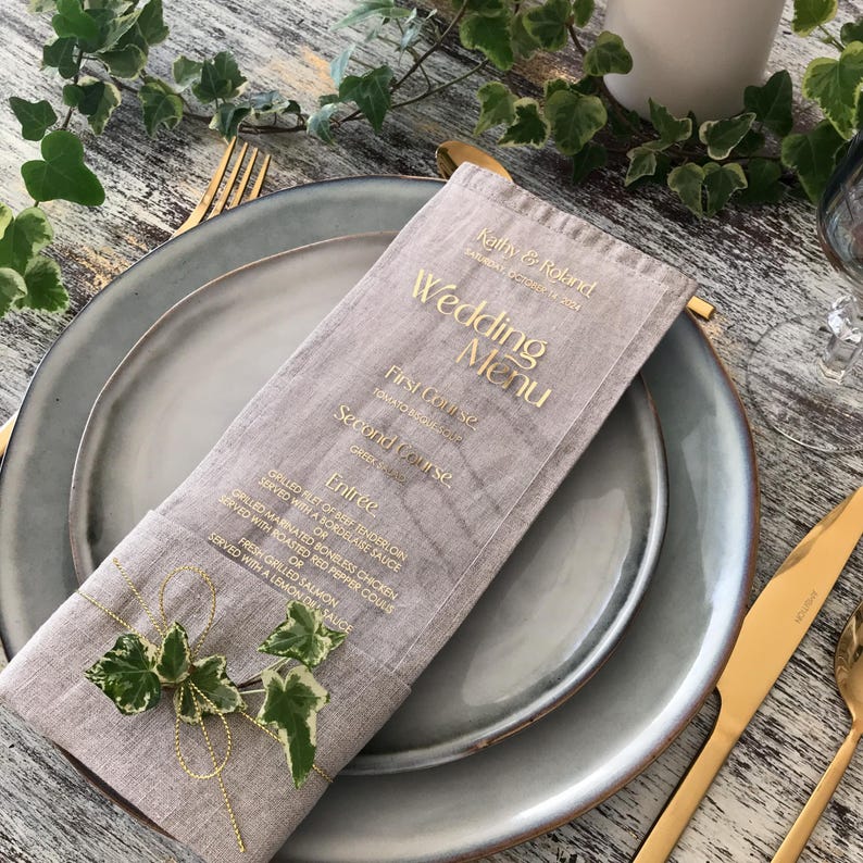 Op de afbeelding: Een trouwmenu op een grijze plaat met gouden bestek. Het menu staat in een linnen hoes met gouden letters met de tekst "Wedding Menu". Het menu is versierd met een takje groene en witte klimop.