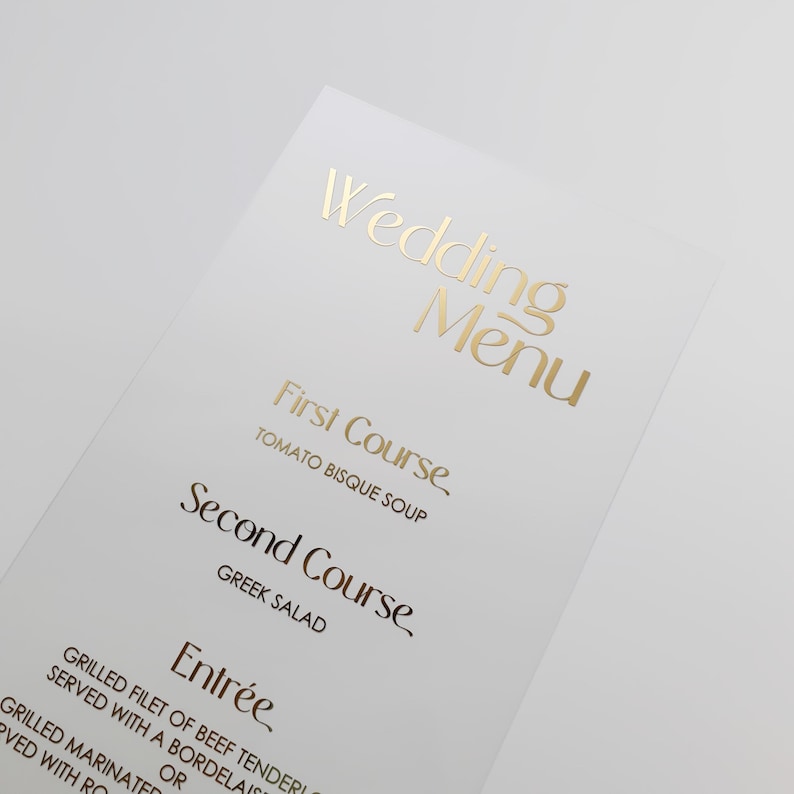 Op de afbeelding: Een wit trouwmenu met gouden folie letters. De tekst luidt "Wedding Menu" bovenaan, gevolgd door de gangen: "First Course, Tomato Bisque Soup", "Second Course, Greek Salad" en "Entr&eacute;e".
