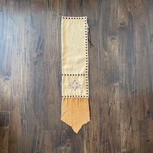 May include: A beige suede bag with brown fringe and a decorative embroidered design. The bag has a brown leather strap and is decorated with brown stitching.