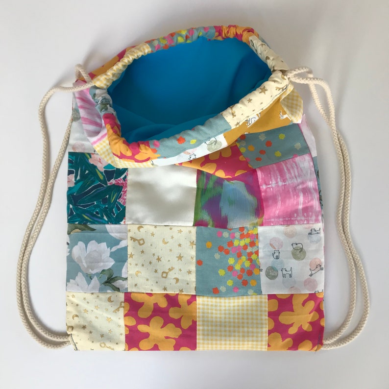 Może przedstawiać: Worek sznurkowy w patchworku z niebieską podszewką. Worek wykonany jest z r&oacute;żnych tkanin z kwiatowymi, geometrycznymi i abstrakcyjnymi wzorami w odcieniach r&oacute;żowego, ż&oacute;łtego, zielonego i niebieskiego.