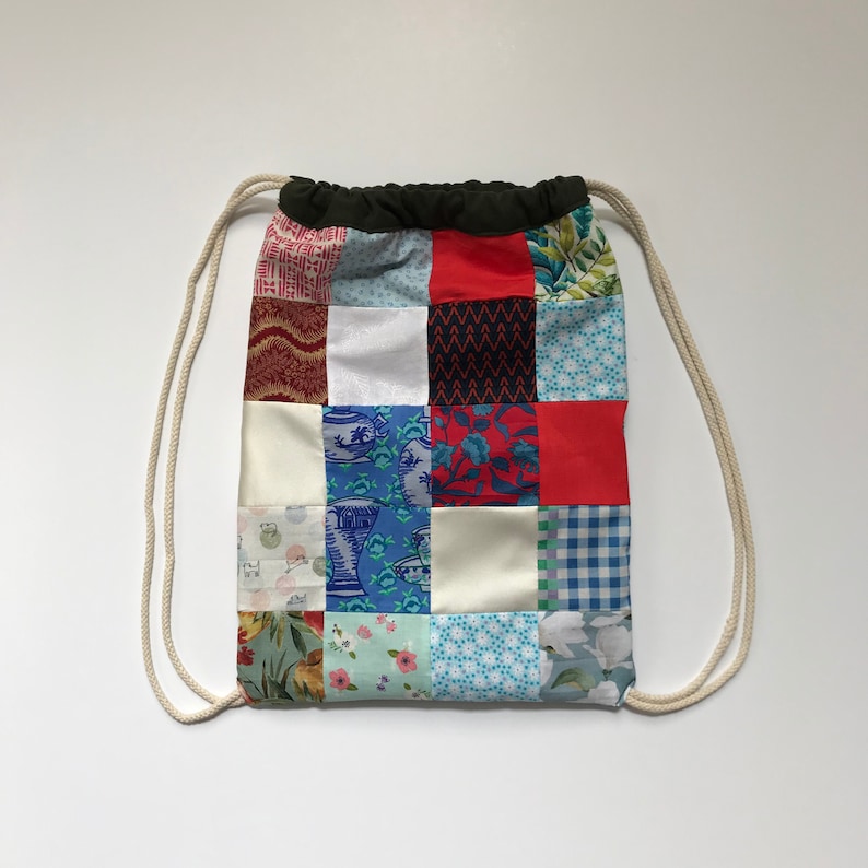 Op de afbeelding: Een patchwork trekkoordtas met verschillende kleurrijke stoffen, waaronder rood, blauw, groen en wit. De tas heeft een wit trekkoord en een zwarte bovenkant.