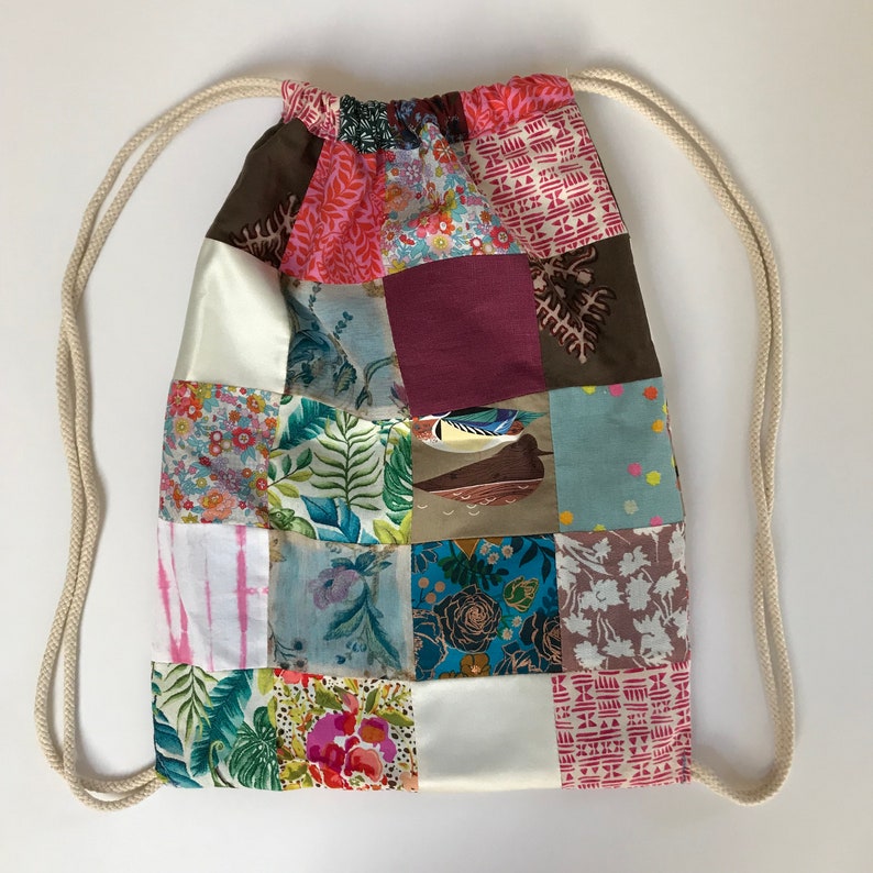 May include: A patchwork drawstring bag with a white cord. The bag is made of various fabrics in shades of pink, brown, blue, green, and white. The bag has a square shape and is made of different patterns.