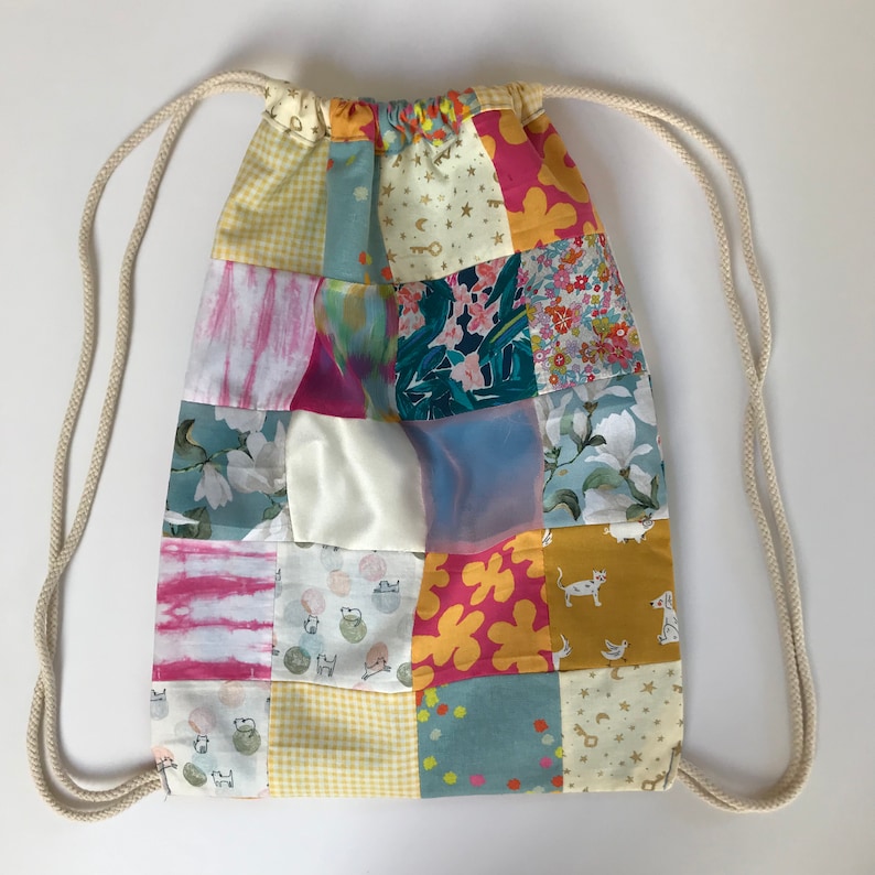 Może przedstawiać: Torba sznurkowa w patchworku z r&oacute;żnymi wzorami kwiatowymi i geometrycznymi w odcieniach r&oacute;żowego, ż&oacute;łtego, niebieskiego i zielonego. Torba ma biały sznurek i jest wykonana z bawełnianej tkaniny.