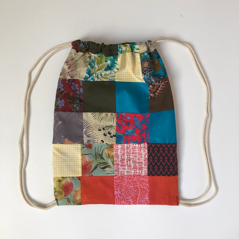 May include: A patchwork drawstring bag featuring a variety of patterned fabric squares in shades of blue, green, red, and orange. The bag has a cream-coloured drawstring closure.
