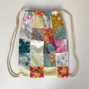 May include: A patchwork drawstring bag with a variety of colorful fabrics, including pink, yellow, blue, and green. The bag has a white drawstring and is made of cotton.