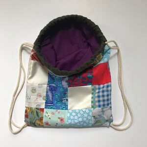 Op de afbeelding: Een patchwork trekkoordtas met een paarse voering. De tas is gemaakt van verschillende stoffen in tinten blauw, rood, groen en wit.