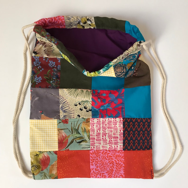 May include: A patchwork drawstring bag with a variety of colourful fabric squares. The bag has a purple interior and cream-coloured rope drawstrings. The fabrics include floral, geometric, and solid patterns in shades of red, blue, green, and yellow.