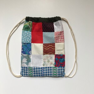 Op de afbeelding: Een patchwork-rugzak met een verscheidenheid aan kleurrijke stoffen, waaronder rood, blauw, wit en groen. De rugzak heeft een wit trekkoord en een donkergroene bovenkant.