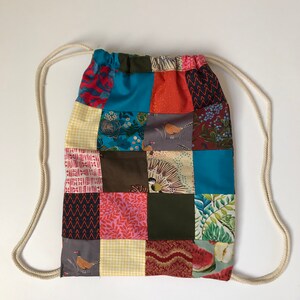 May include: A patchwork drawstring bag featuring squares of patterned fabric in blue, red, orange, green, and brown. It has a cream-coloured drawstring closure. The bag is set against a white background.
