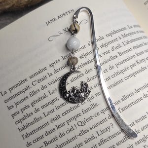 Può includere: Un segnalibro in metallo argentato con un ciondolo a forma di luna crescente e castello, ornato da perline bianche e beige. Il segnalibro è appoggiato su un libro aperto con testo francese.