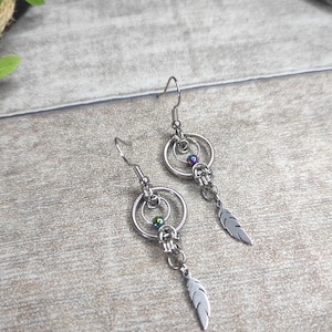 Może przedstawiać: Srebrne kolczyki wiszące z zawieszką w kształcie pióra. Każdy kolczyk posiada dwa połączone okrągłe kółka, małe koraliki i zwisujące pióro. Kolczyki są prezentowane na jasnoszarej drewnianej powierzchni.