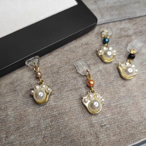 Op de afbeelding: Vier paar kattenpootvormige oorbellen. Elke oorbel heeft een goudkleurig pootafdrukontwerp met imitatieparels. De oorbellen hebben een doorzichtige plastic pin en een kleine accentkraal in verschillende kleuren.