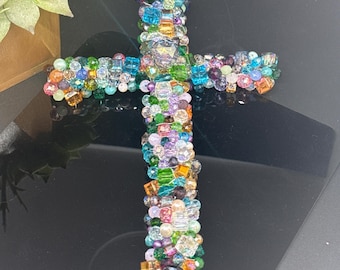 May include: A colorful beaded cross made with a variety of glass beads in shades of green, blue, purple, pink, and orange. The cross is a religious symbol and a decorative piece.