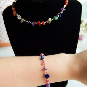 May include: A gold chain necklace and bracelet with colorful gemstone chips. The necklace is on a black display stand and the bracelet is on a person's wrist.