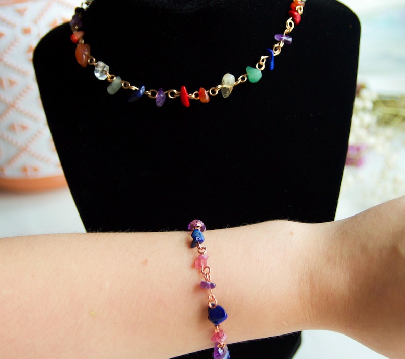 May include: A gold chain necklace and bracelet with colorful gemstone chips. The necklace is on a black display stand and the bracelet is on a person's wrist.