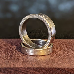 May include: Two silver rings made from coins, stacked on top of each other. The top ring has the words "LIBERTY" and "IN GOD WE TRUST" engraved on it. The bottom ring has the words "E PLURIBUS UNUM" and "AMERICA" engraved on it.
