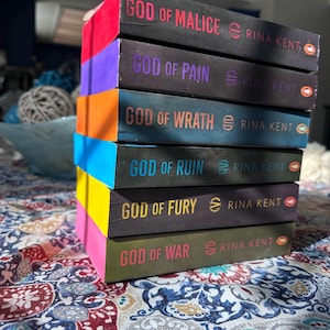 A complete 6-book set of Rina Kent's Legacy of Gods series featuring custom sprayed edges. The book edges are airbrushed in a solid rainbow gradient of red, purple, orange, blue, yellow, and pink to perfectly match the original covers. The books are stacked on a white shelf in a minimalist "BookTok" aesthetic style.