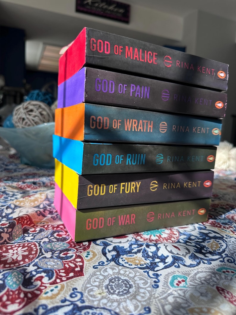 A complete 6-book set of Rina Kent's Legacy of Gods series featuring custom sprayed edges. The book edges are airbrushed in a solid rainbow gradient of red, purple, orange, blue, yellow, and pink to perfectly match the original covers. The books are stacked on a white shelf in a minimalist "BookTok" aesthetic style.