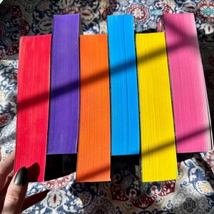 A complete 6-book set of Rina Kent's Legacy of Gods series featuring custom sprayed edges. The book edges are airbrushed in a solid rainbow gradient of red, purple, orange, blue, yellow, and pink to perfectly match the original covers. The books are stacked on a white shelf in a minimalist "BookTok" aesthetic style.