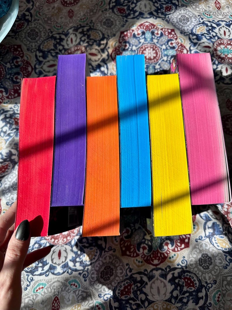 A complete 6-book set of Rina Kent's Legacy of Gods series featuring custom sprayed edges. The book edges are airbrushed in a solid rainbow gradient of red, purple, orange, blue, yellow, and pink to perfectly match the original covers. The books are stacked on a white shelf in a minimalist "BookTok" aesthetic style.