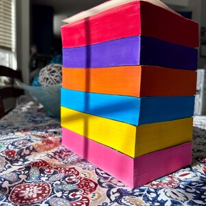 A complete 6-book set of Rina Kent's Legacy of Gods series featuring custom sprayed edges. The book edges are airbrushed in a solid rainbow gradient of red, purple, orange, blue, yellow, and pink to perfectly match the original covers. The books are stacked on a white shelf in a minimalist "BookTok" aesthetic style.