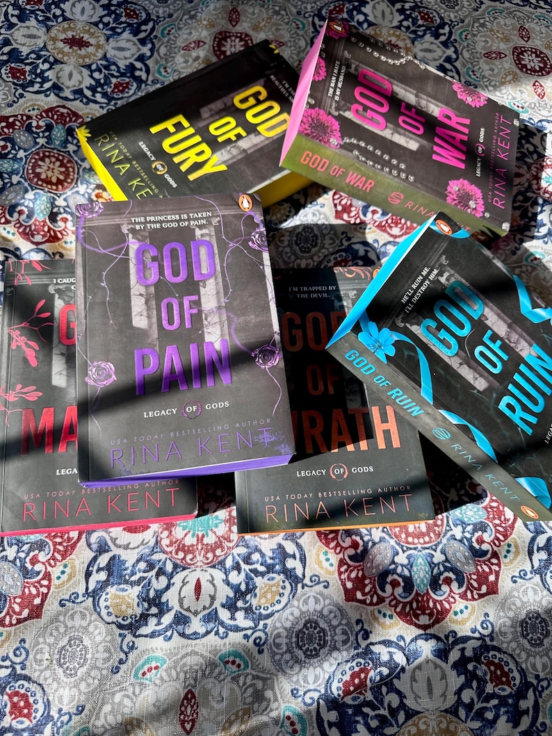 A complete 6-book set of Rina Kent's Legacy of Gods series featuring custom sprayed edges. The book edges are airbrushed in a solid rainbow gradient of red, purple, orange, blue, yellow, and pink to perfectly match the original covers. The books are stacked on a white shelf in a minimalist "BookTok" aesthetic style.