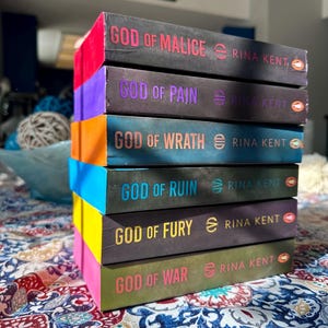 May include: A stack of six books with colorful spines. The titles include "God of Malice," "God of Pain," "God of Wrath," "God of Ruin," "God of Fury," and "God of War" by Rina Kent. The books are stacked on a patterned surface.
